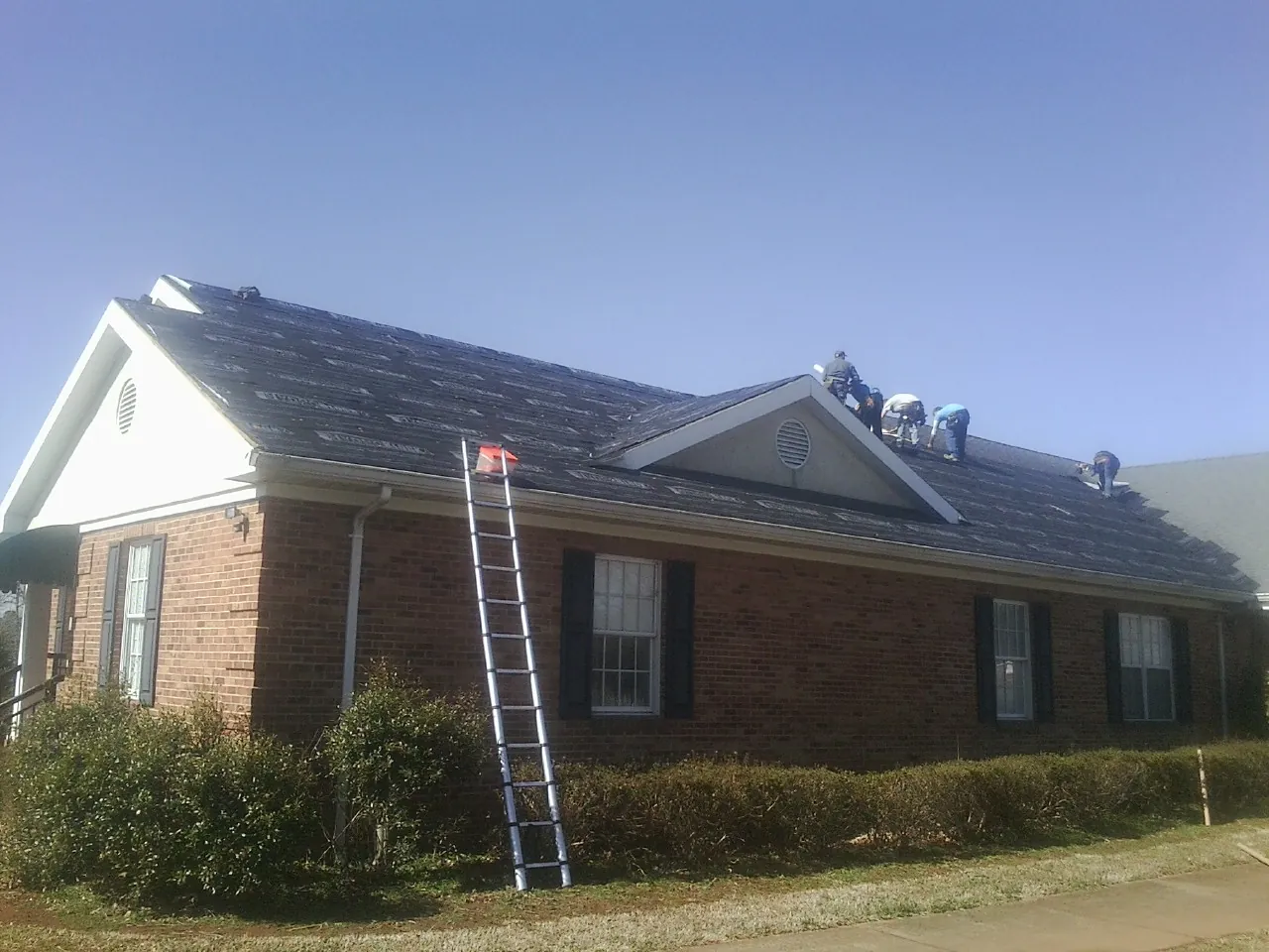 Crew tearing shingles off a brick ranch roof with a ladder set — Emergency Roof Repair work by our crew in Fort Payne