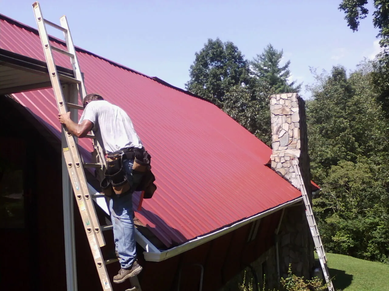 Roofer on a ladder beside a red metal roof and stone chimney — Soffit & Fascia Repair by Roofing Brothers in Fort Payne